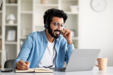 A smiling man looks at his laptop whilst making notes in a notebook. 