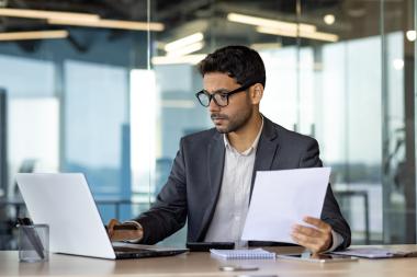 A man compares information on a laptop screen against a piece of paper.